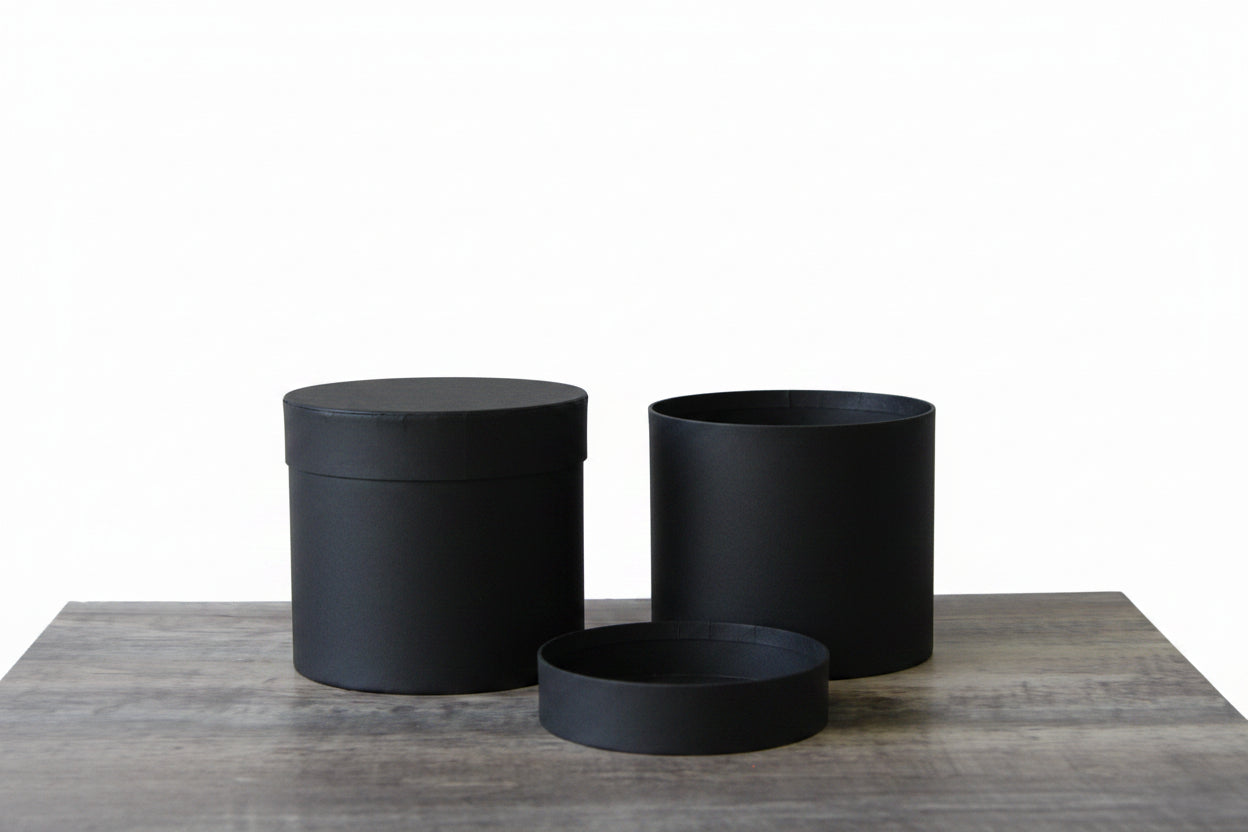 Two matte black cylindrical containers or lids placed side by side on a rustic wooden surface, with soft shadows cast on a light wall behind them.