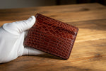 Brown crocodile skin wallet held by a gloved hand on a neutral background