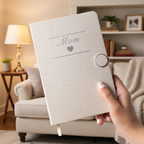 Beige notebook with 'Mom' and a heart embroidered on it, held by a hand against a white background.