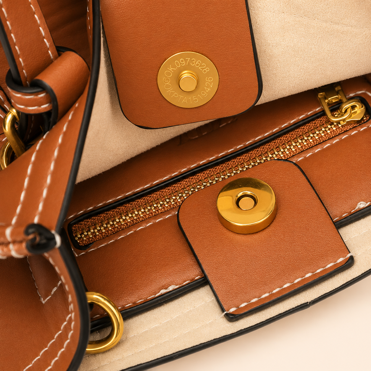 Close-up of a brown leather handbag interior showing a zipper, snap button closure, white contrast stitching, and gold-tone hardware on a light beige background.