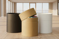 Three cylindrical storage boxes or containers on a tabletop: one matte black, one kraft brown with its lid partly open showing a stack inside, and one matte white. Soft shadow overlays are visible on the wall behind them.