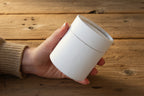 Hand holding a white cylindrical container against a brown background
